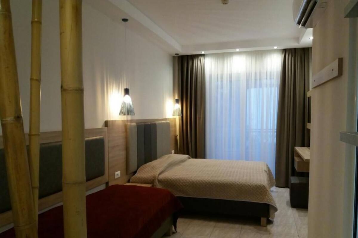 Una habitación de hotel con cama, mesita de noche, lámpara y ventana con cortinas.