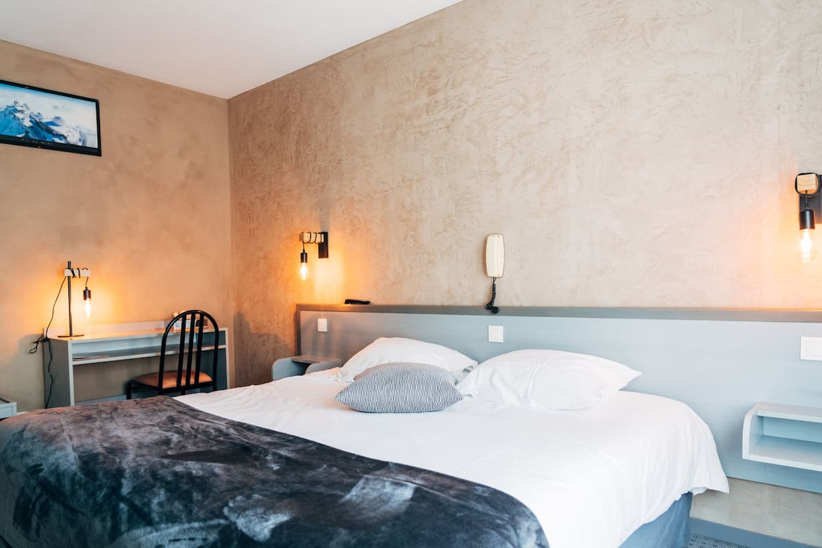 Ein Hotelzimmer mit Bett, Schreibtisch und Stuhl. An der Wand sind Wandleuchten und ein Fernseher angebracht.