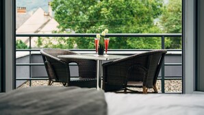 Un balcón con sillones de mimbre y una mesa, un florero con flores, y vistas a árboles y edificios.