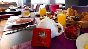 Una mesa de desayuno con un calentador de té, tazas de té, un vaso de jugo de naranja, croissants y una bolsita de té KUSMI.