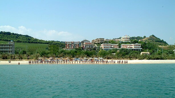 A coastal area with a beach, buildings, and a hilly background. - Residence Azzurro (Massignano)