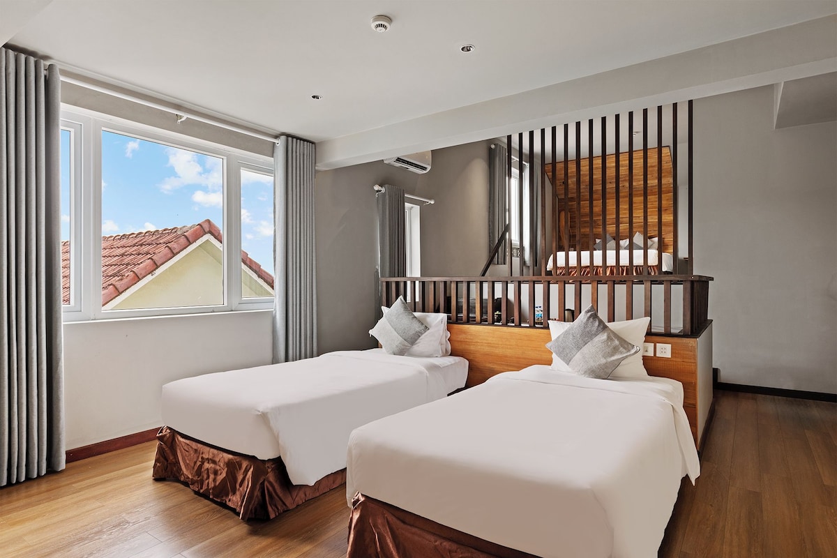 A hotel room with two beds, a large window, a wooden headboard, and a view of the roof.