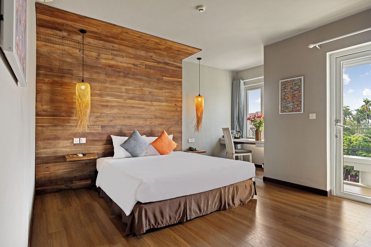 A modern bedroom with a wooden headboard, a large bed with white bedding, two pendant lights, and a view of the outdoors through a sliding glass door.