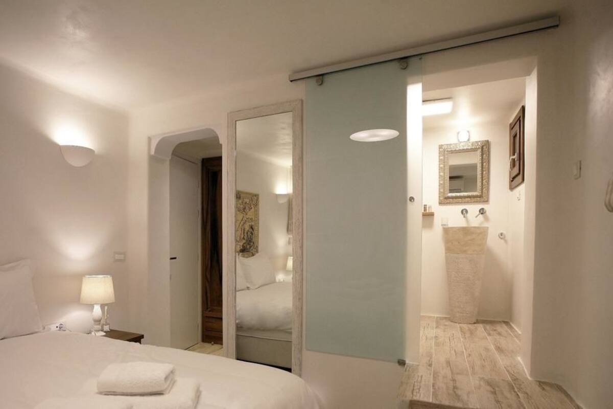 A hotel room with a bed, a nightstand, a lamp, a mirror, and a bathroom with a sink and mirror.