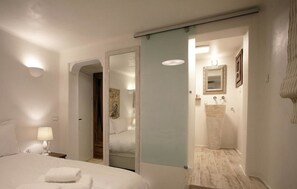A hotel room with a bed, a nightstand, a lamp, a mirror, and a bathroom with a sink and mirror. - Anagram Hotel (Mykonos)