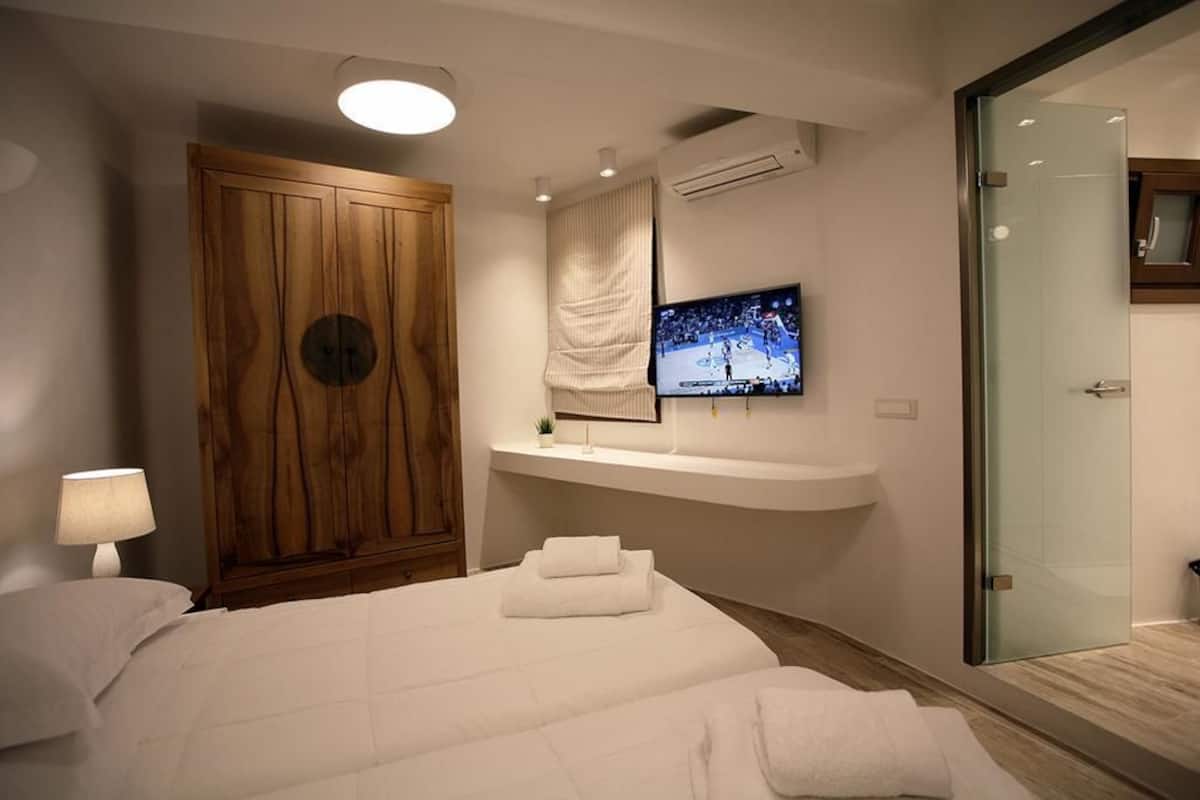 A modern hotel room with a large bed, a TV mounted on a wall, a wooden wardrobe, and a glass shower enclosure.