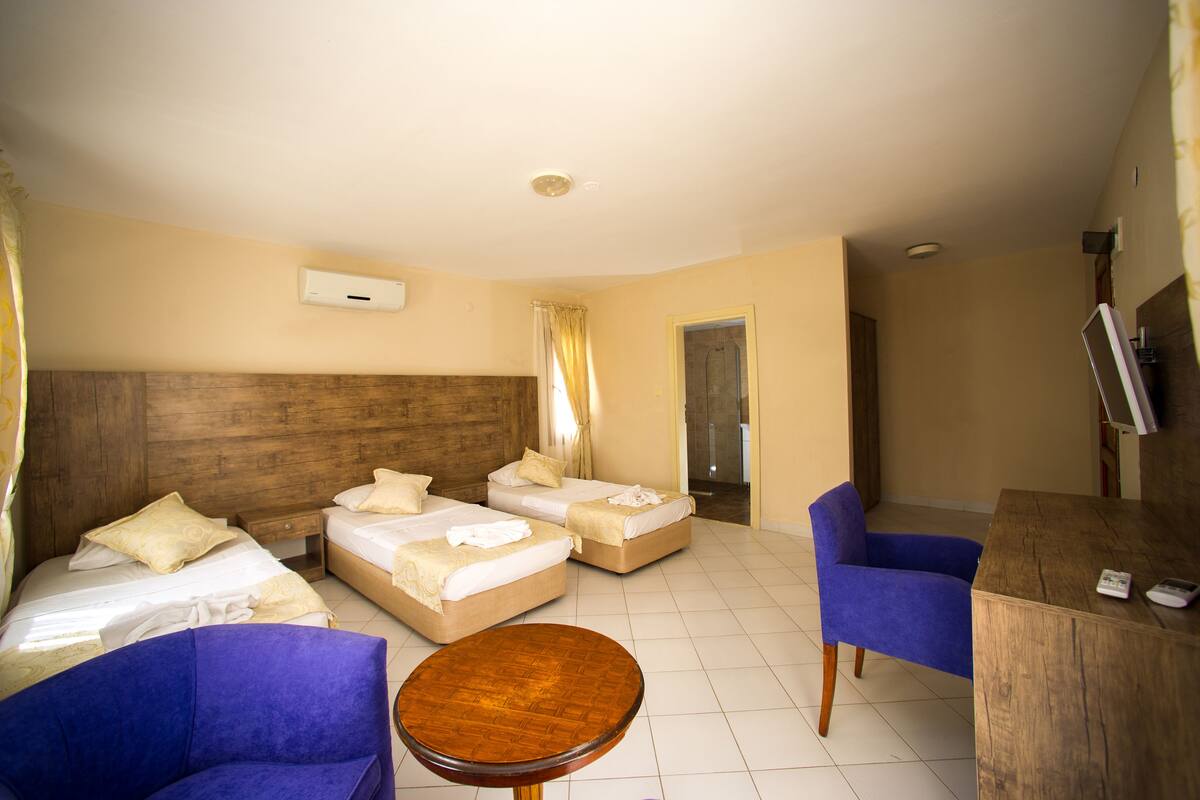 Quarto de hotel com duas camas, uma cabeceira de madeira, uma cadeira azul, uma mesa de madeira e uma televisão sobre um suporte de madeira.