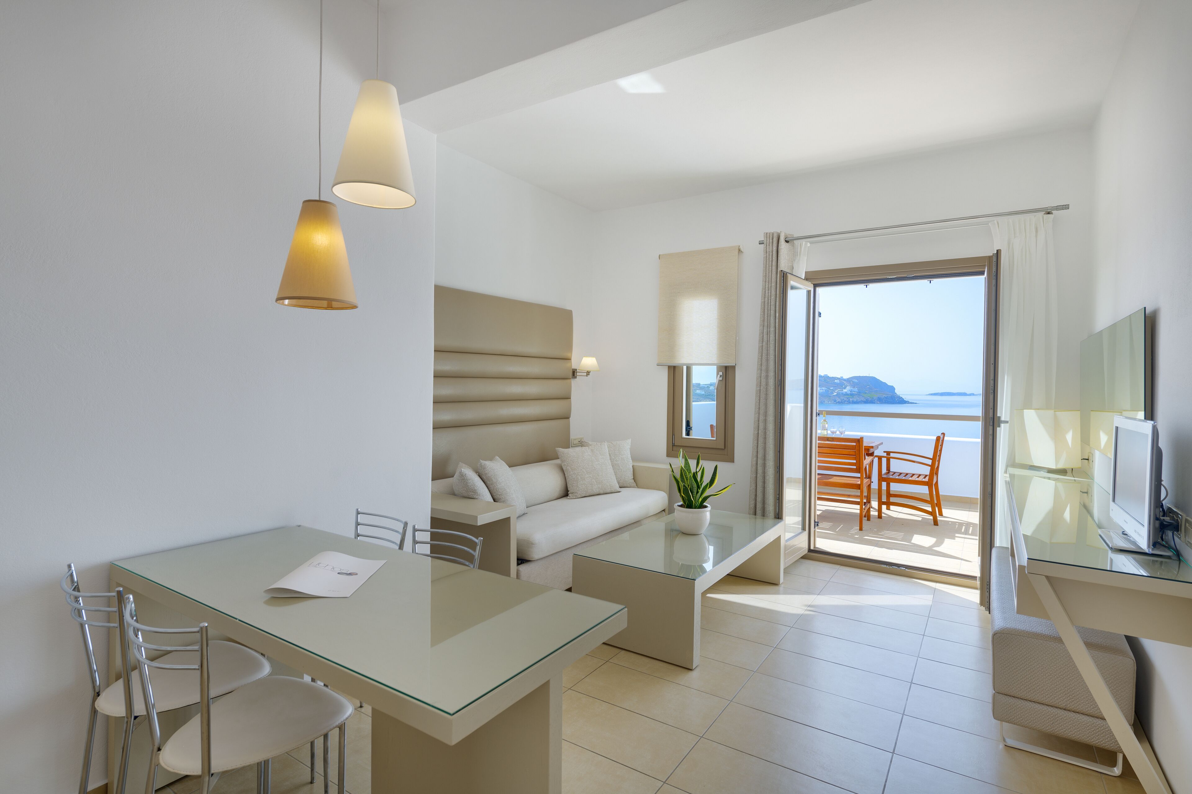 A modern hotel room with a glass dining table, a sofa, a TV, and a balcony with a view of the sea.