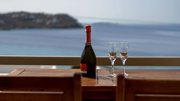 Uma garrafa de vinho e dois copos sobre uma mesa de madeira, com vista para o mar e uma ilha ao longe.