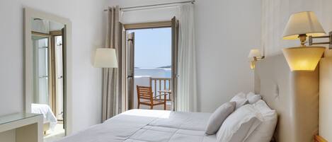 A hotel room with a bed, a desk, a chair, and a view of the sea through an open door.