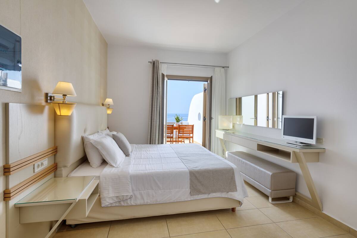 A hotel room with a bed, bedside table, TV, and a view of the sea through a window.