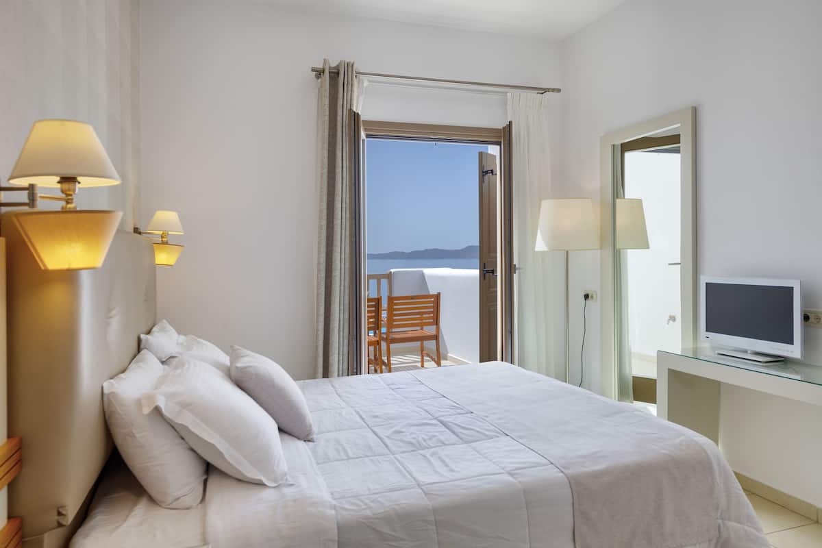 A hotel room with a bed, a chair, a desk with a TV, and a view of the sea.