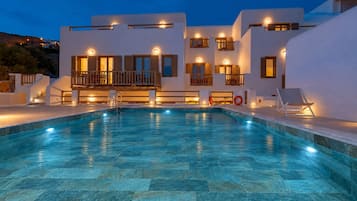 Ein Schwimmbad mit einer Terrasse aus Steinplatten, umgeben von einem weißen Gebäude mit Balkonen und warmer Beleuchtung.