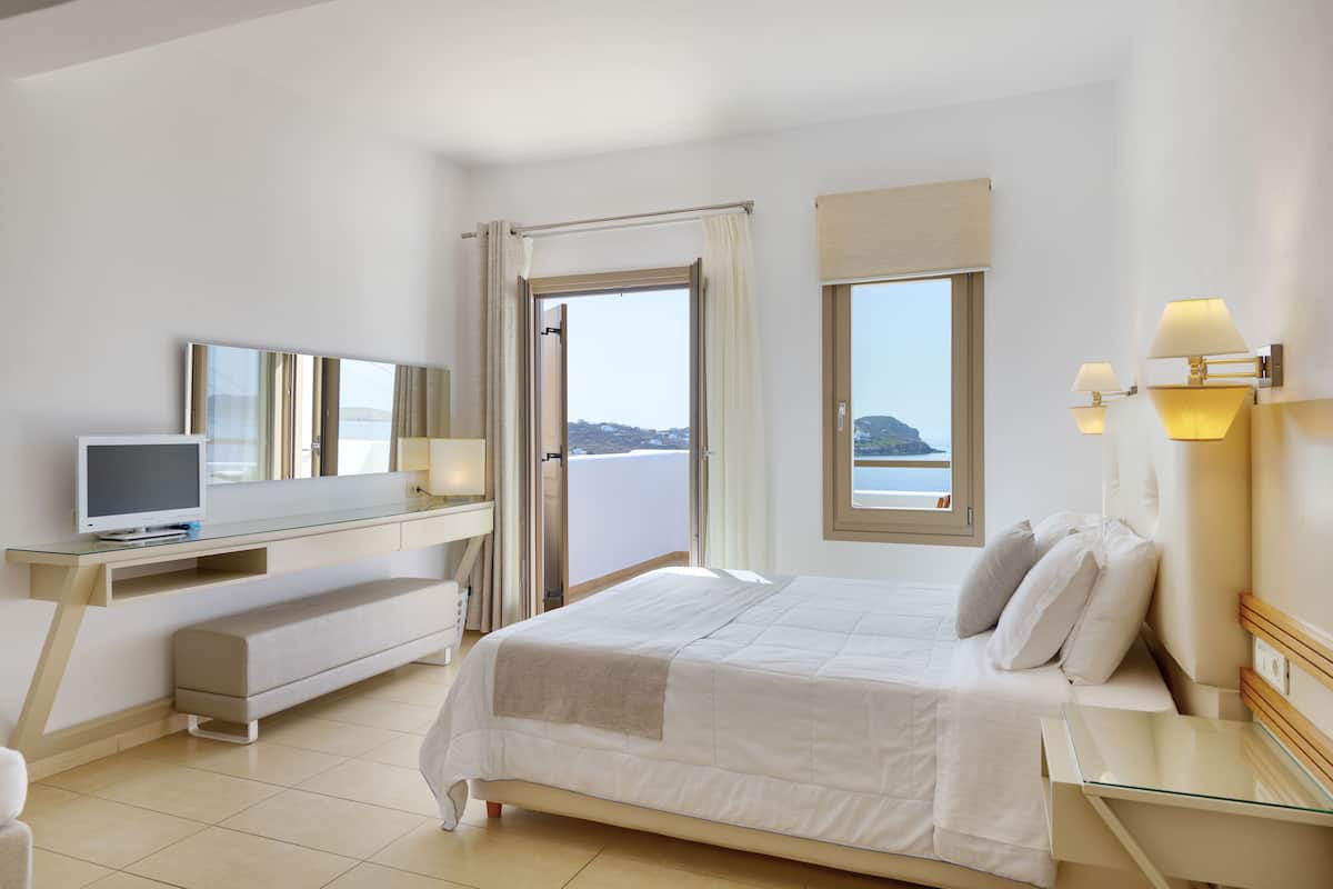 A modern hotel room with a bed, a desk, a TV, and a large window offering a view of the sea.