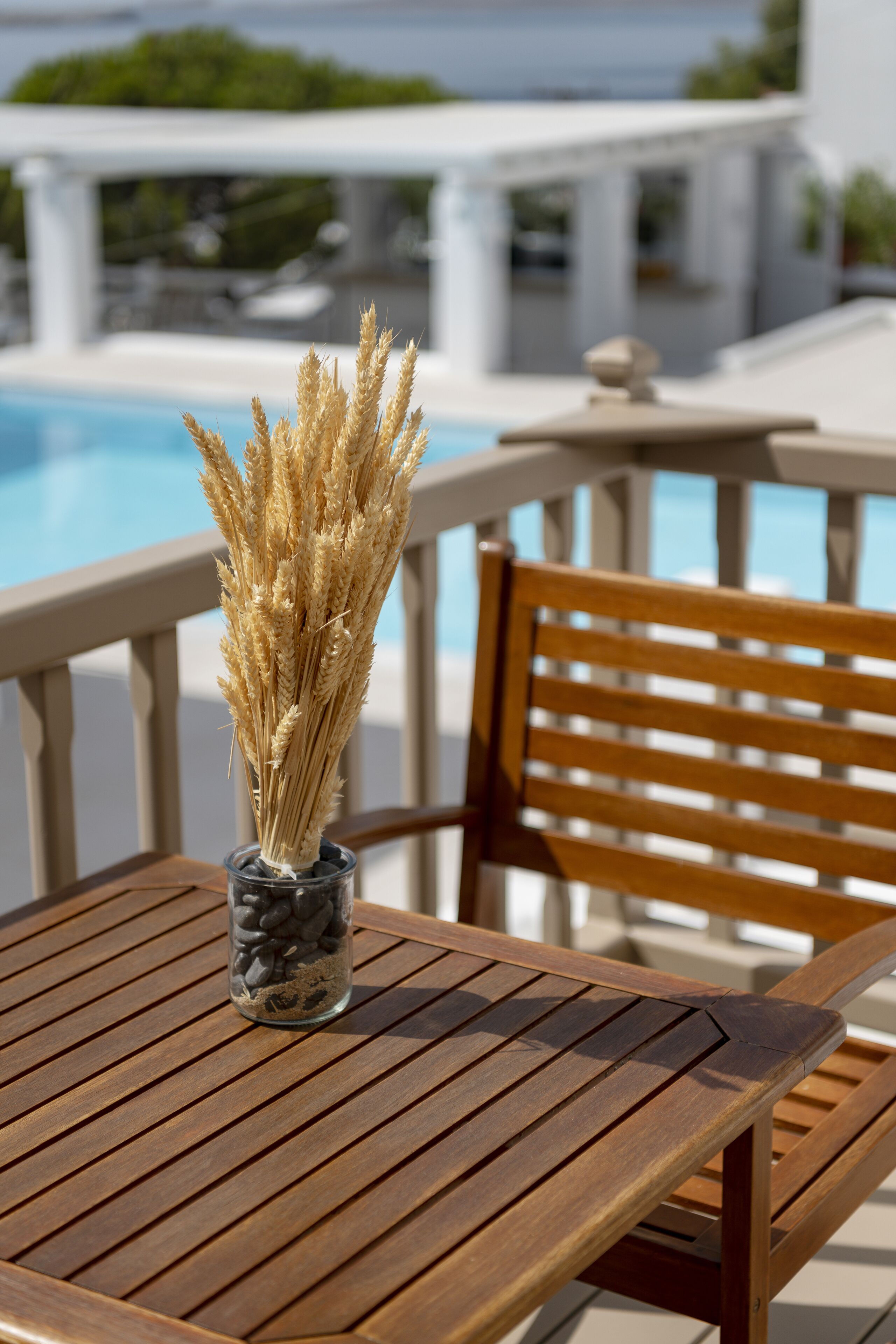 Una mesa de madera con un jarrón de espigas de trigo, una silla de madera y una piscina al fondo.