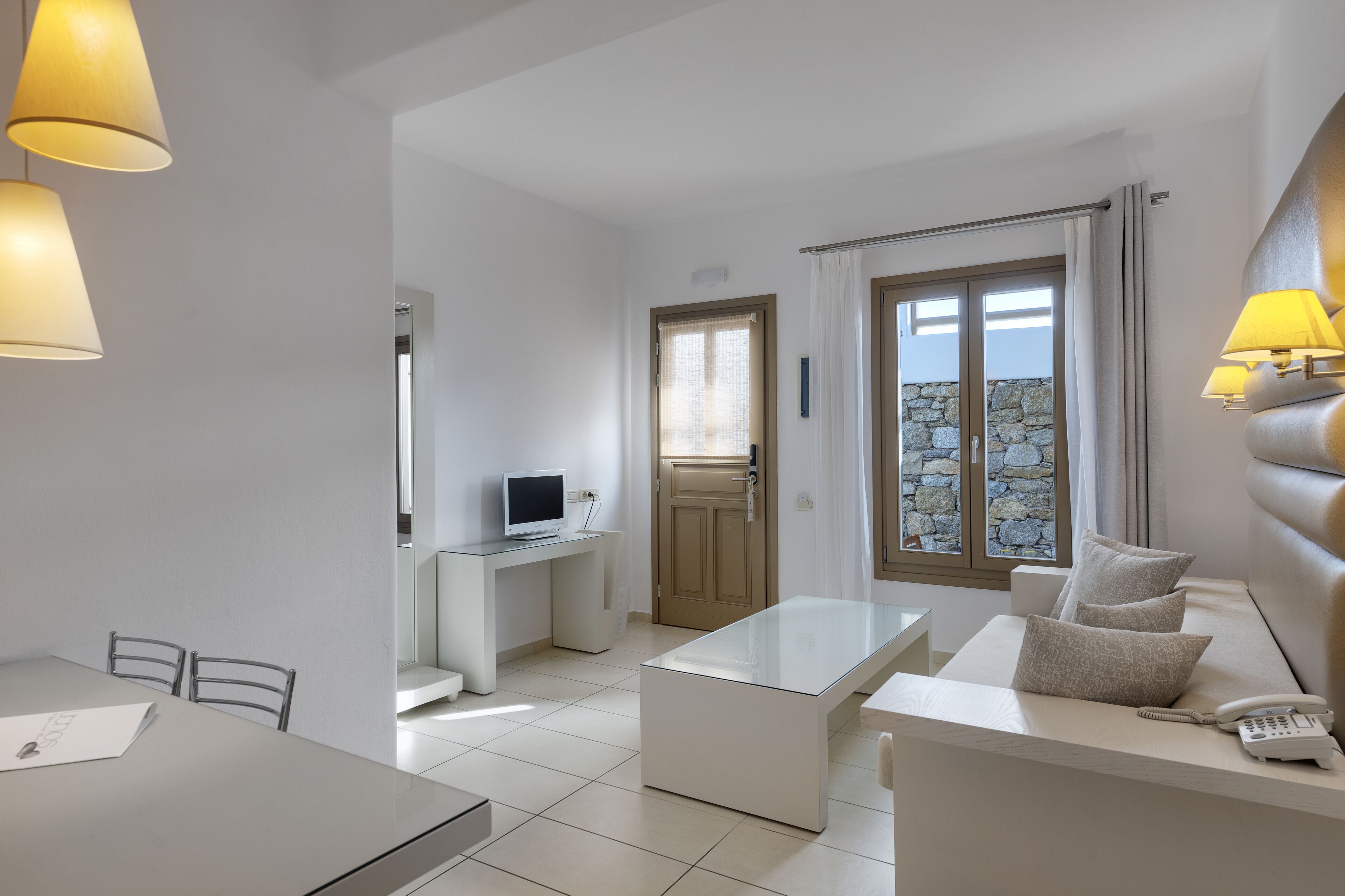 A modern hotel room with a dining area, a small desk with a computer, a sofa with cushions, and a stone wall visible through the window.