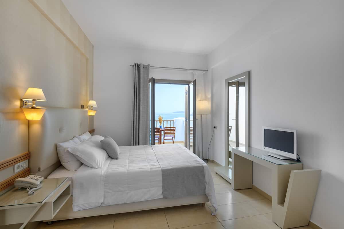 A hotel room with a bed, bedside lamps, a desk with a TV, and a view of the sea through a balcony door.