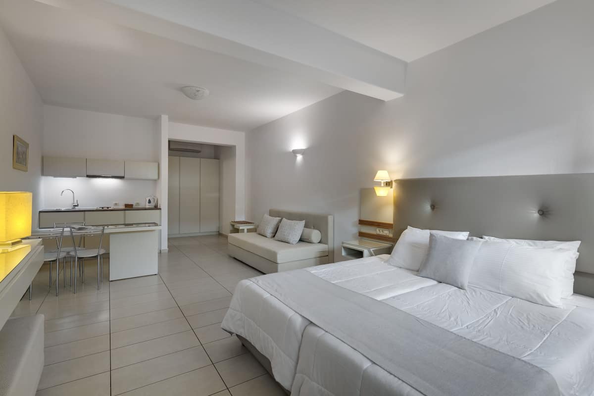 A modern hotel room with a large bed, a desk with a chair, a sofa, and a kitchenette.