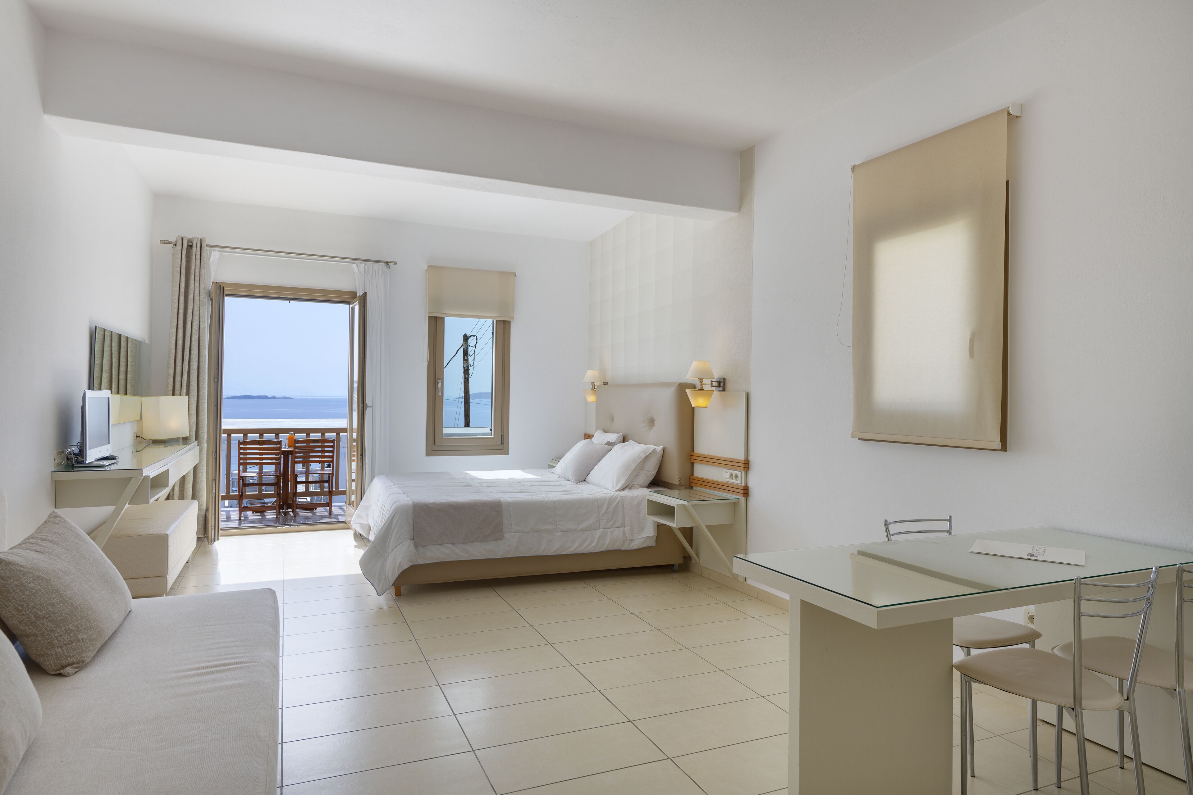 A hotel room with a bed, a desk, a chair, a sofa, a balcony with a view, and a large window.