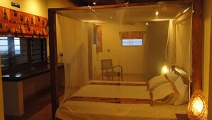 A room with a bed covered by a mosquito net, a chair, a small table with a lamp, and a window with curtains. - Goodlife Residence (Bujumbura)