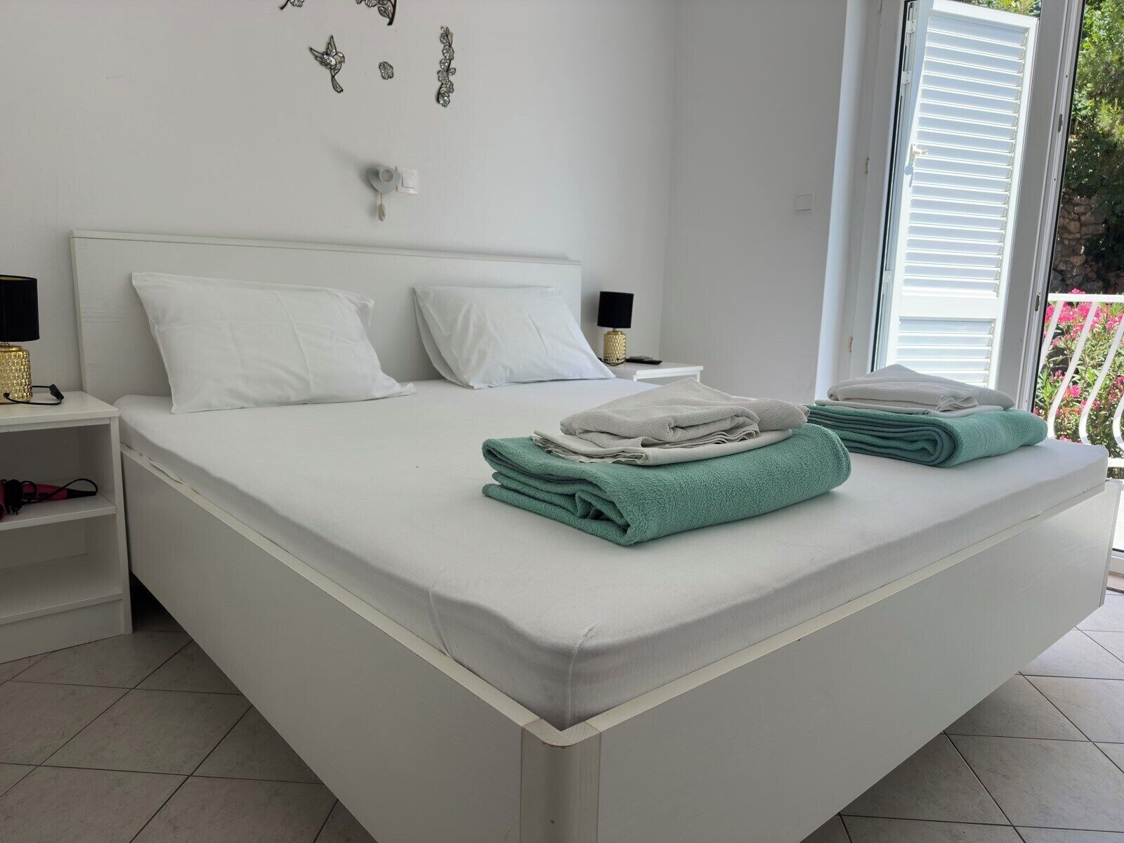 Un dormitorio con cama, dos mesitas de noche blancas, una ventana con persianas y un balcón con barandilla.