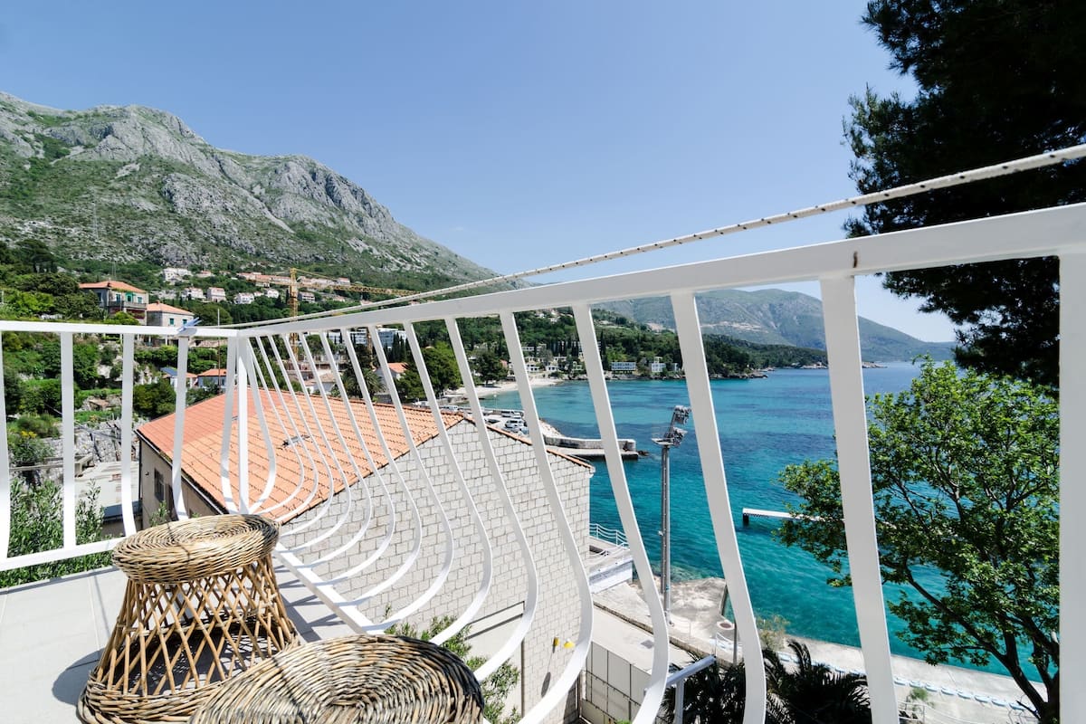 A balcony with wicker furniture overlooking a coastal area with clear blue waters and a mountainous backdrop.