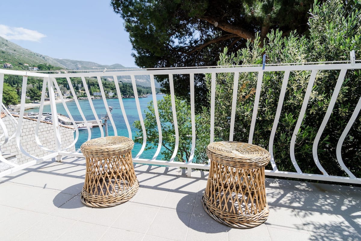 A balcony with wicker stools, a white railing, and a view of a coastal area with boats and greenery.