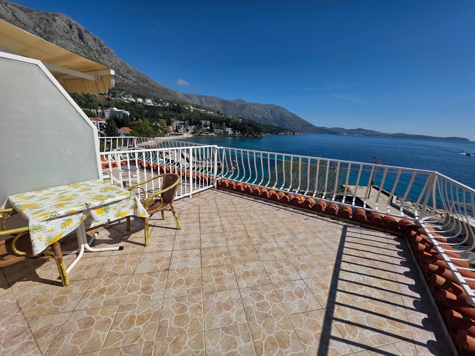 A balcony with a table and chairs, a railing, and a view of the sea and mountains.