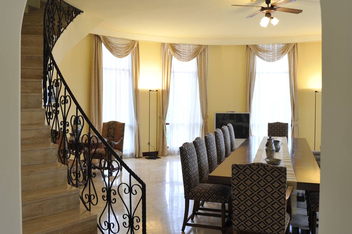 A dining area with a long table, patterned chairs, a staircase with ornate railings, and a ceiling fan.