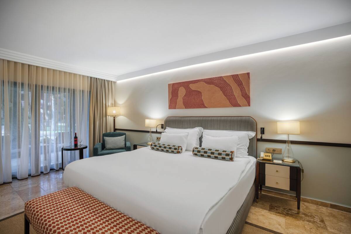 A hotel room with a large bed, two bedside tables, a chair, a bench, and a view of the outdoors.