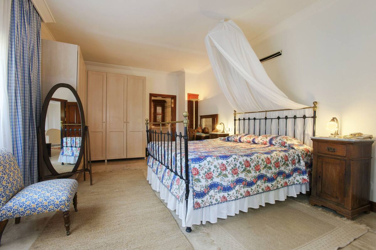 A bedroom with a canopy bed, a wooden wardrobe, a mirror, a chair, and a bedside table.