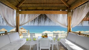 A covered outdoor seating area with white cushions and a table with fruit, overlooking a beach. - Martı Myra - All Inclusive (Kemer)