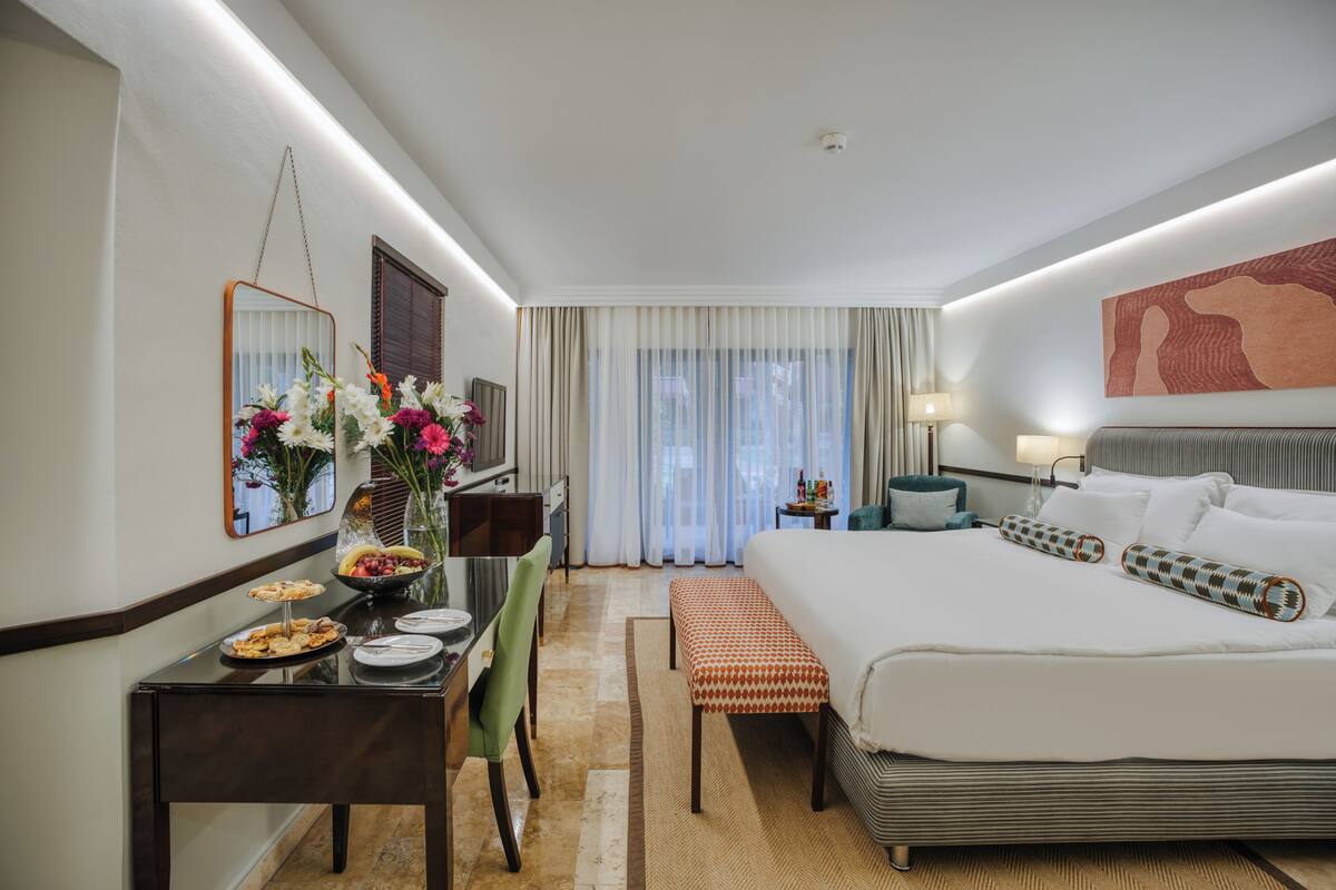 A hotel room with a bed, bedside table, lamp, and a view of the outdoors.