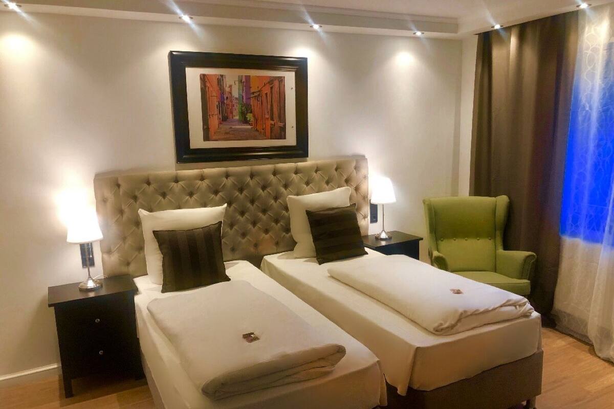 A hotel room with two beds, a green armchair, a framed artwork on the wall, and a window with curtains.