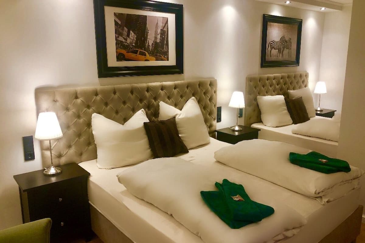 A hotel room with two beds, each with a tufted headboard, white bedding, and a green towel. There are framed pictures on the wall and a lamp on each bedside table.
