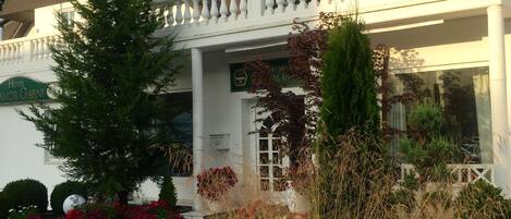 Ein Hotel mit weißer Fassade, einem Balkon mit filigraner Treppe und einem Garten mit Bänken und blühenden Pflanzen.