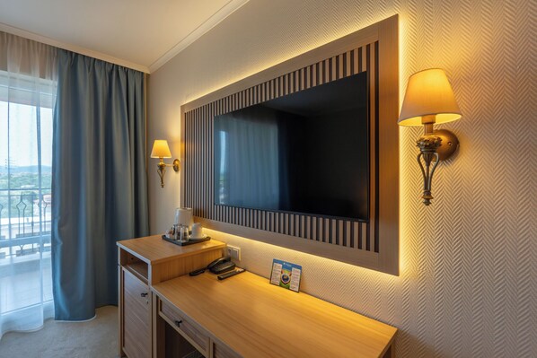A hotel room with a wooden desk, a flat-screen TV, two wall-mounted lamps, a window with curtains, and a view of the outdoors. - DIT Majestic Beach Resort (Nessebar)