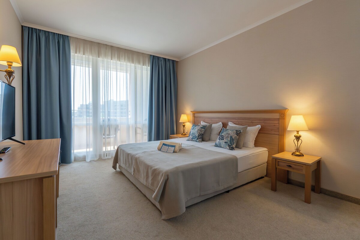 Een hotelkamer met een bed, nachtkastjes, een bureau en uitzicht op de stad.