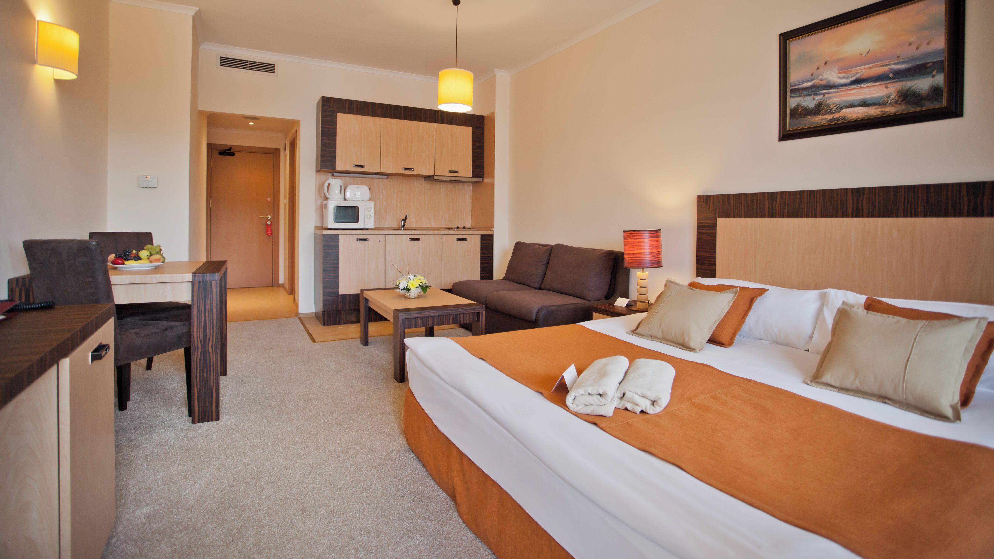 A hotel room with a bed, a desk with a chair, a sofa, a small kitchenette, and a painting on the wall.
