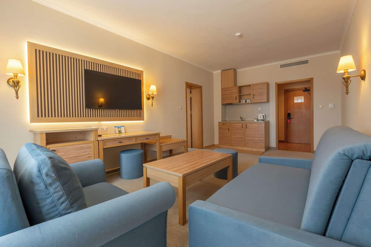 A modern living room with a flat-screen TV, a wooden coffee table, blue sofas, and a kitchen area in the background.