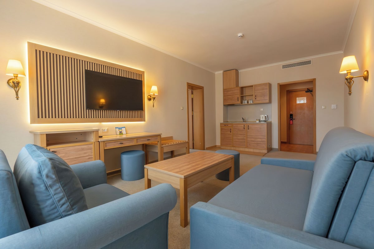 Un salon moderne avec un téléviseur à écran plat, une table basse en bois, des canapés bleus et un coin cuisine à l’arrière-plan.