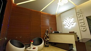Un área de recepción moderna de hotel, con un mostrador de recepción, dos sillas negras y un elemento decorativo en la pared.