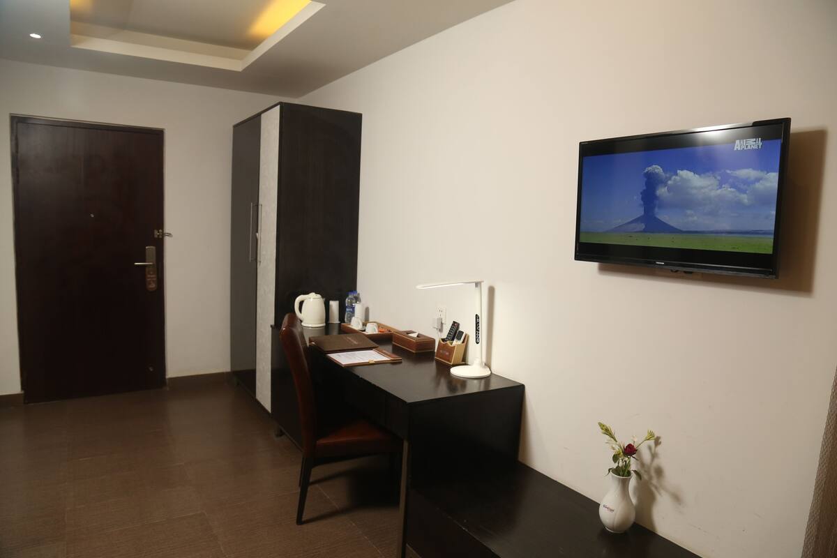 Habitación de hotel con un escritorio de madera oscura, una silla, un pequeño jarrón blanco con flores, un televisor en la pared y una puerta.