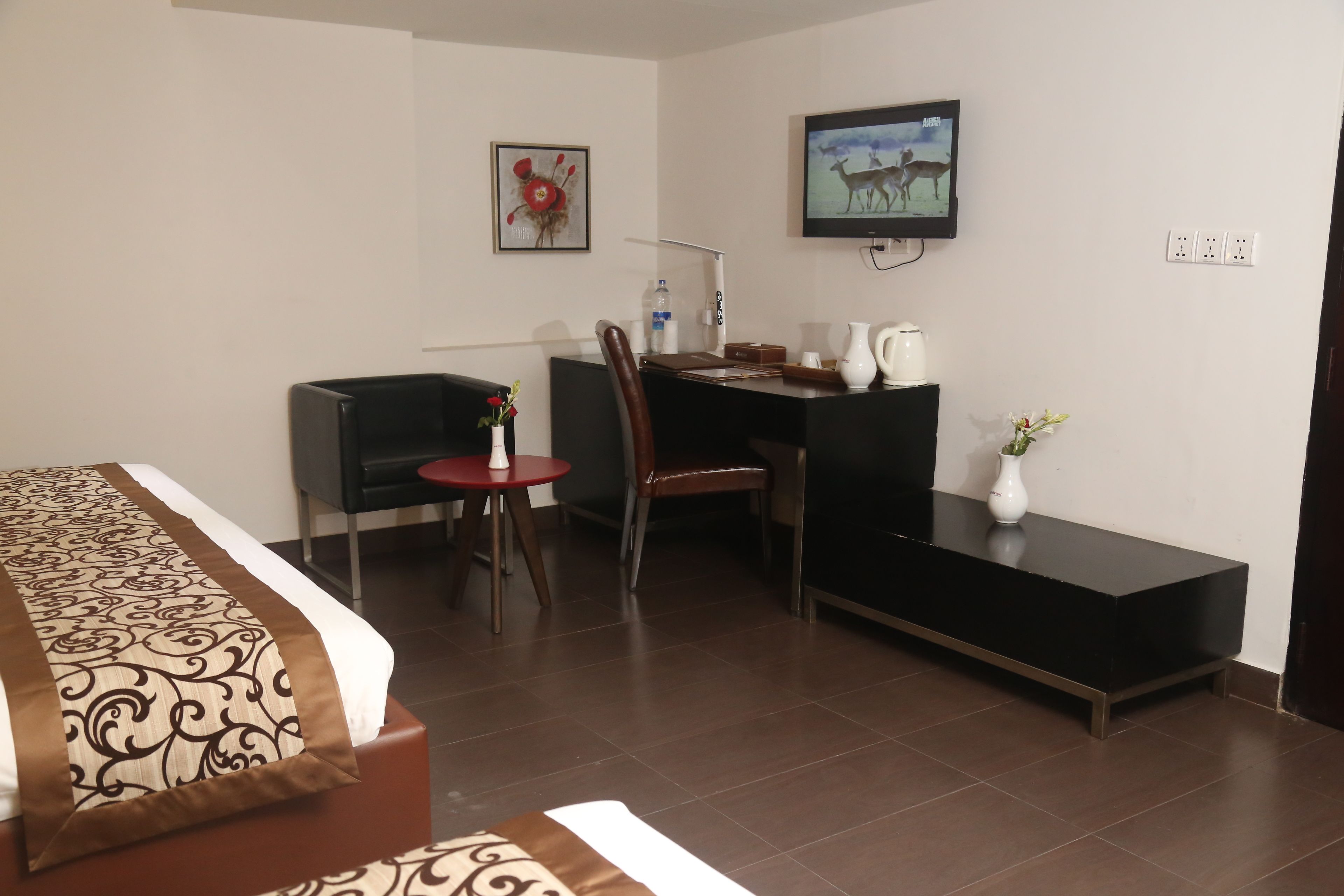A hotel room with a bed, a desk, a chair, a small table, a TV, and a wall-mounted picture.