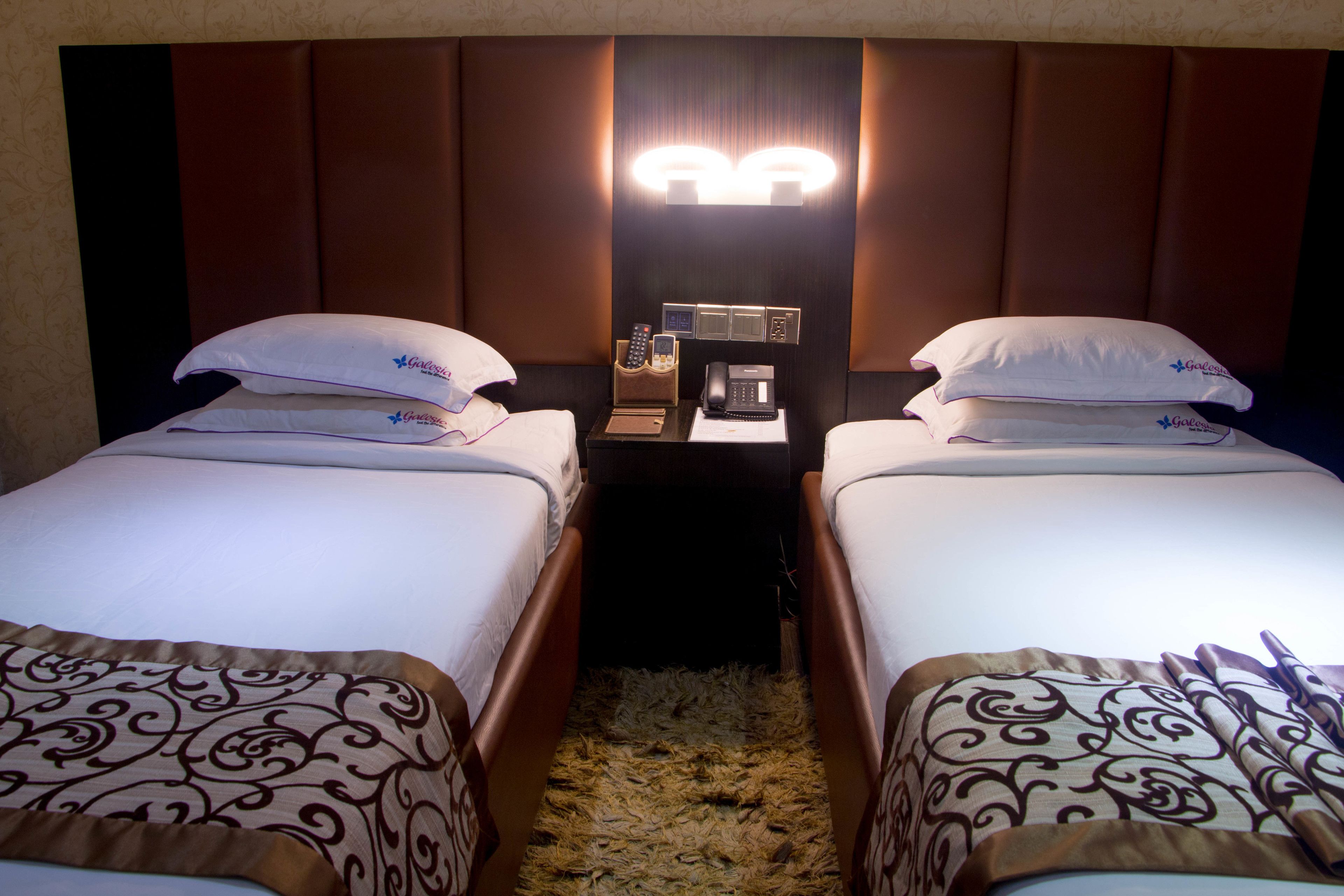 Two single beds with patterned bedspreads, a nightstand with a telephone and a lamp, and a wall-mounted light fixture.