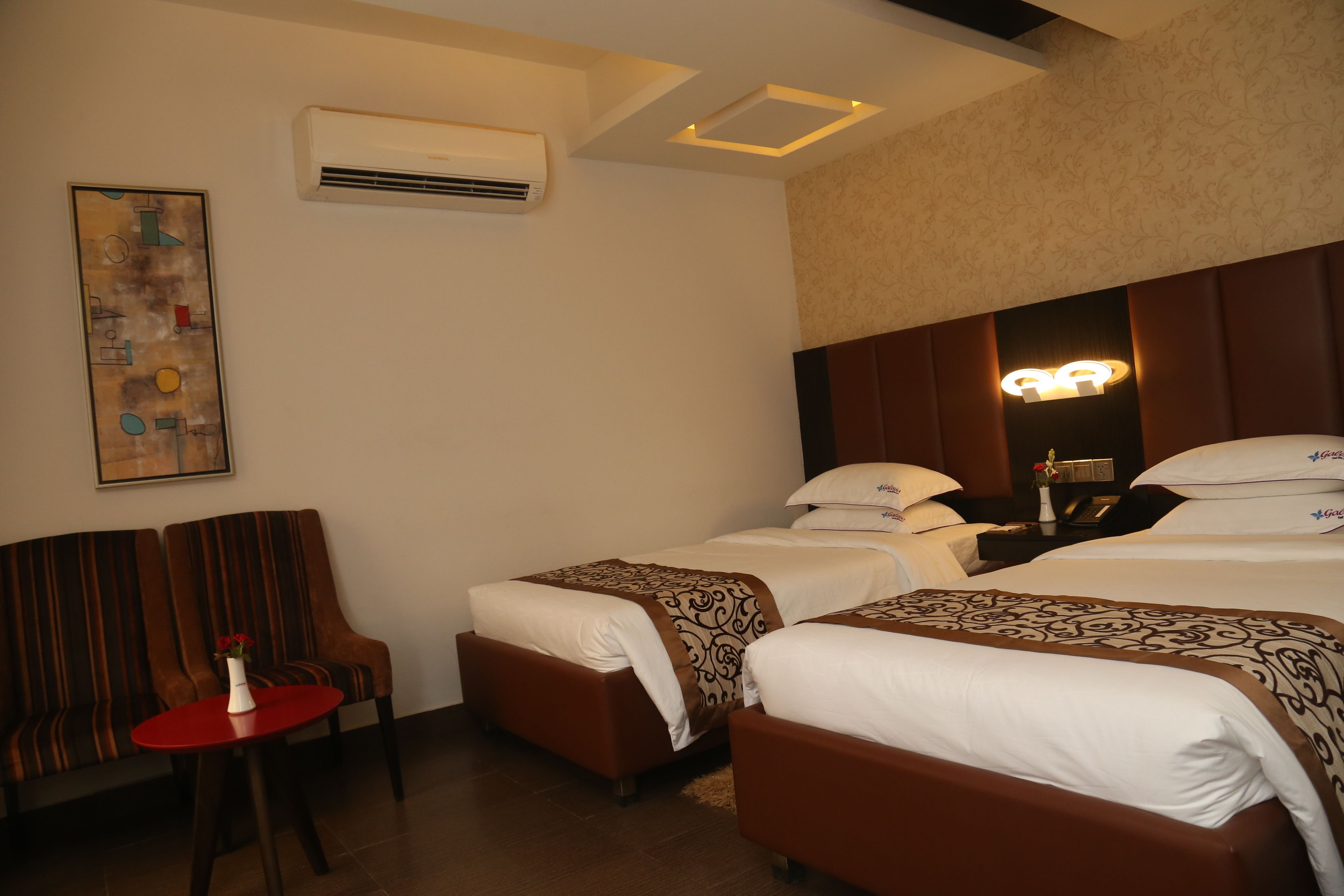 Habitación de hotel con dos camas, una mesa roja, una silla, aire acondicionado y una obra de arte colgada en la pared.