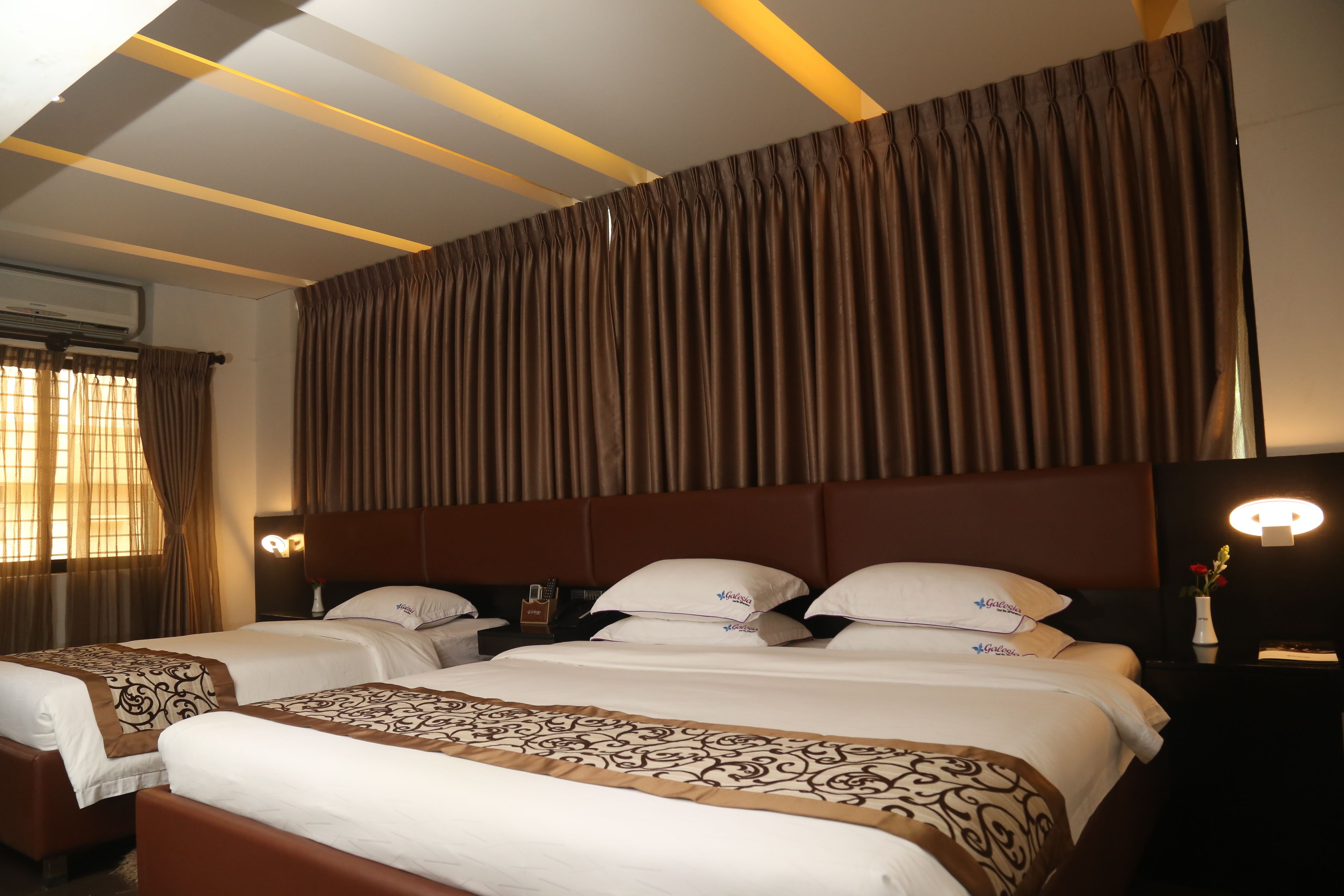 A hotel room with two beds, a large window with curtains, a wall-mounted air conditioner, and a bedside table with a lamp and a vase of flowers.
