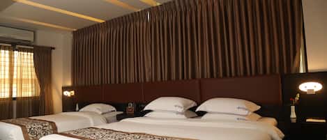 A hotel room with two beds, a large window with curtains, a wall-mounted air conditioner, and a bedside table with a lamp and a vase of flowers.