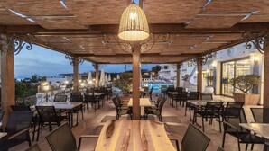 Un área de comedor al aire libre con mesas y sillas de madera, una lámpara colgante y vistas de los edificios y el paisaje circundante.