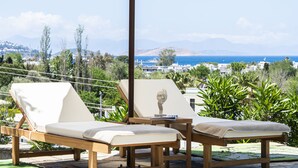 Dos sillones de madera con cojines color beige, una mesita y una escultura sobre ella. El fondo muestra una vista de un pueblo costero con edificios y montañas.
